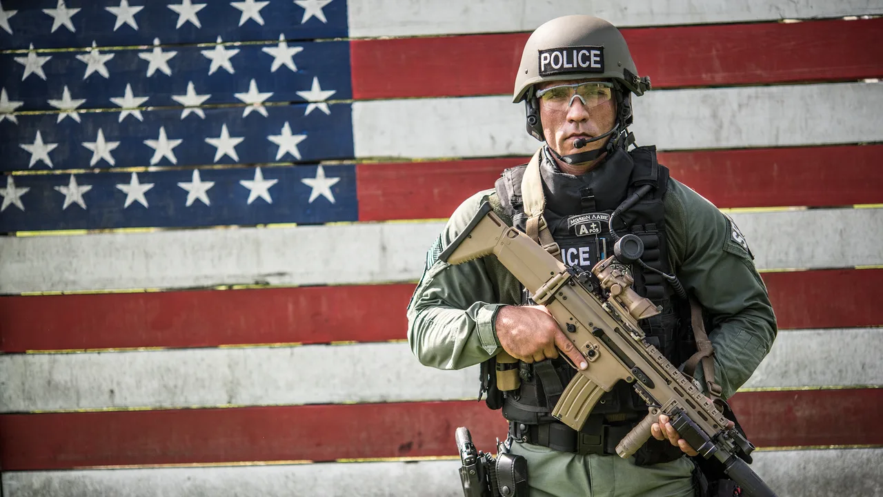 Agente de ICE con equipo táctico frente a una bandera de Estados Unidos