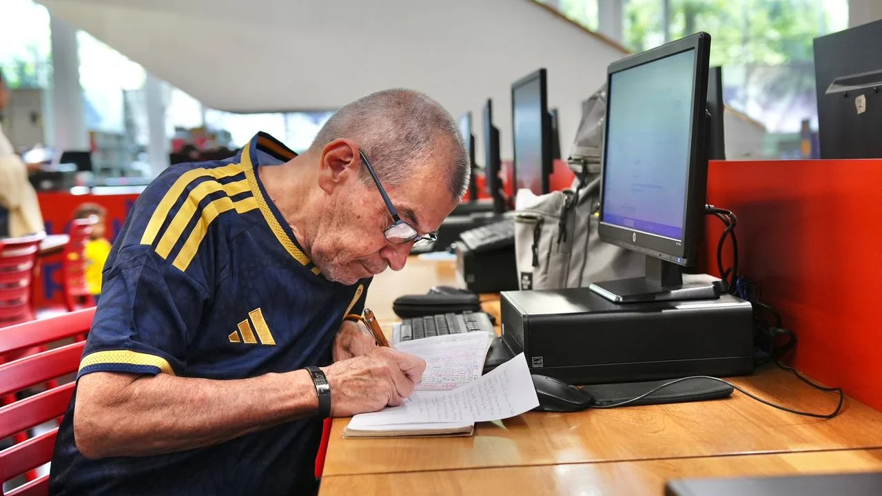 Educación digital gratis llega a los cinco corregimientos de Medellín: así será en las bibliotecas 1 Un hombre escribe en un cuaderno junto a un computador en una biblioteca